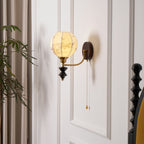 Lit Clérachic globe wall light on a white panelled wall, beside a patterned curtain and the turned post of a black bed.