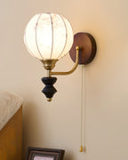 Clérachic globe wall light with a floral print, lit on a cream wall with a pull chain hanging beside a folded striped throw.