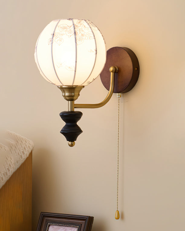 Clérachic globe wall light with a floral print, lit on a cream wall with a pull chain hanging beside a folded striped throw.