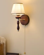 Clérachic wall light with a pleated fabric shade, a dark wood stem and a pull chain, shown on a cream wall.