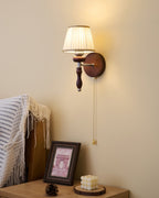 Clérachic wall light with a pleated white shade, lit, mounted above a nightstand with a framed picture and a pine cone.