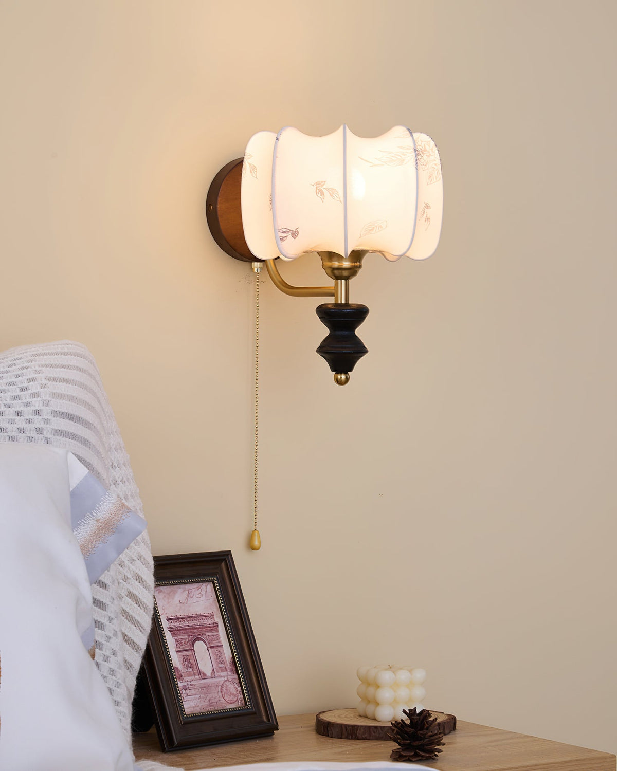 Clérachic wall light with a drum shade and brass pull chain above a nightstand with a small framed print and a candle.