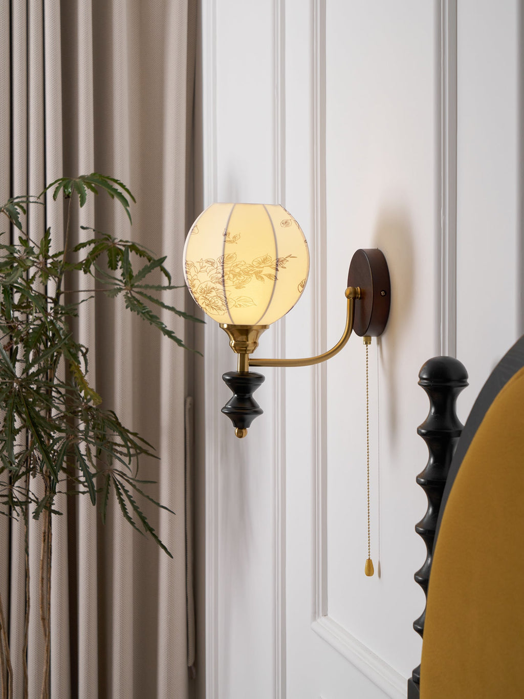 Clérachic globe wall light with a printed shade, lit against a white panelled wall beside a mustard headboard and a leafy plant.