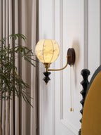 Clérachic globe wall light with a printed shade, lit against a white panelled wall beside a mustard headboard and a leafy plant.