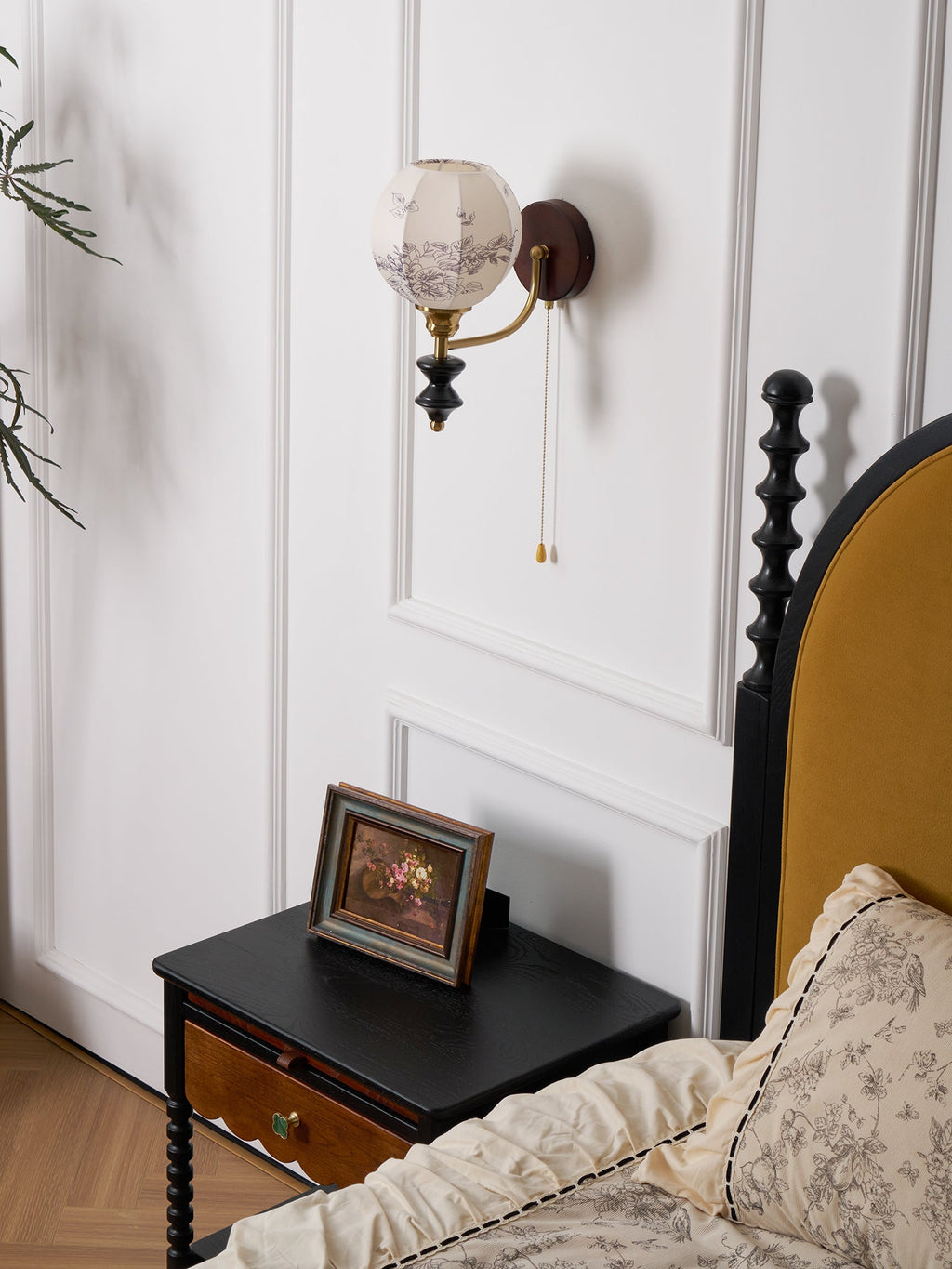 Smaller view of the Clérachic globe wall light above a black nightstand, with a framed picture and a ruffled floral pillow.