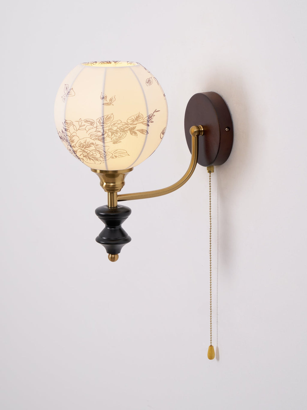 Clérachic globe wall light with a floral-printed fabric shade, brass arm and black wood pendant on a pale wall.