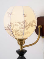 Close view of the floral print on the globe shade of this Clérachic wall light, with its brass cup and arm below.