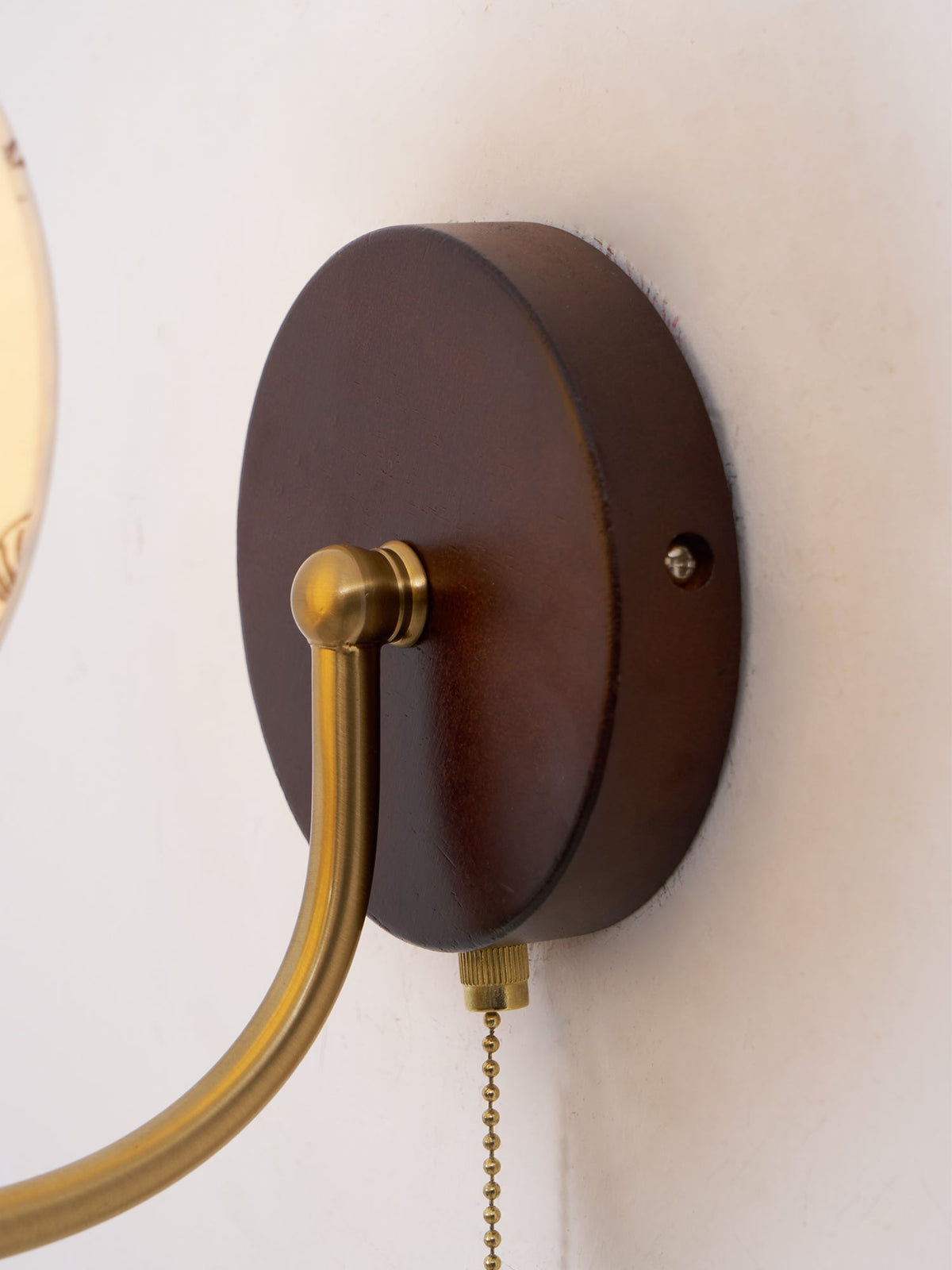 Close view of the round dark wood backplate on this Clérachic wall light, with the brass arm and pull chain leaving it.