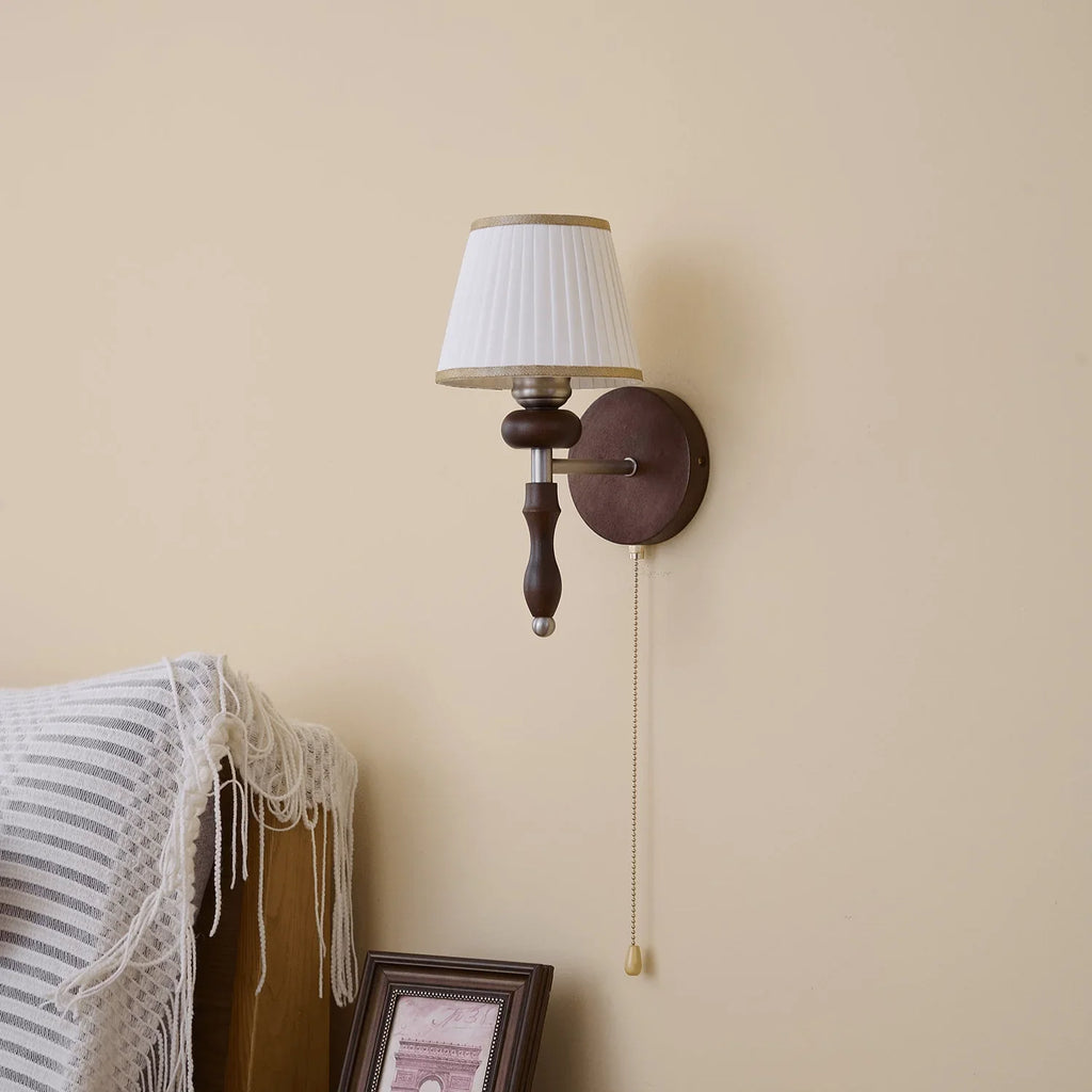 Clérachic wall light with a white pleated shade on a cream wall, with a striped throw and a framed print below.