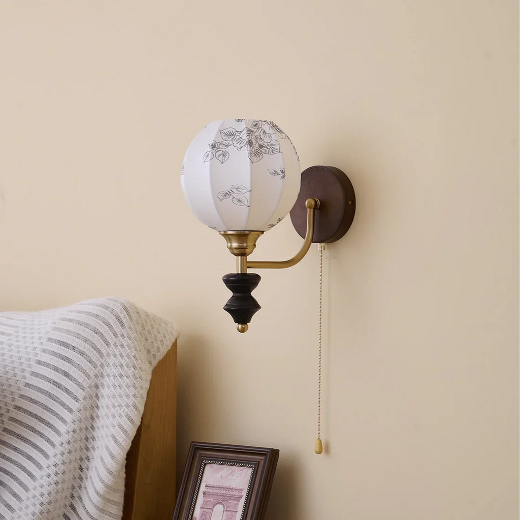 Unlit Clérachic globe wall light with a printed fabric shade on a cream wall, above a striped throw and a framed print.