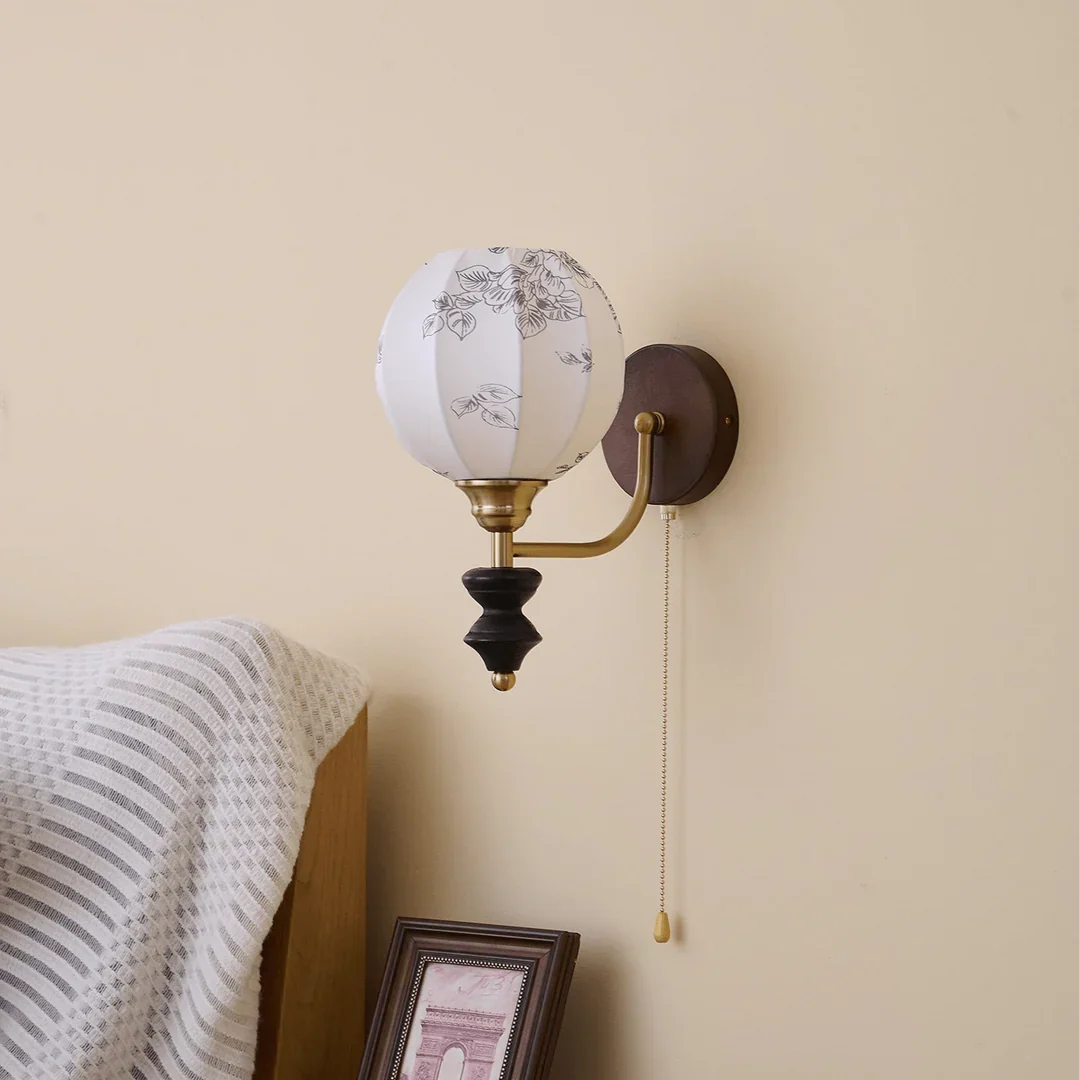Unlit Clérachic globe wall light with a printed fabric shade on a cream wall, above a striped throw and a framed print.