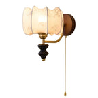 Clérachic wall light with a lit drum shade, a brass arm and a pull chain, on a plain white backdrop.