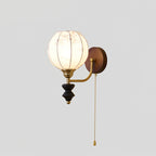 Clérachic wall light with a lit globe shade, a brass arm, a black wood pendant and a pull chain, on a plain grey backdrop.