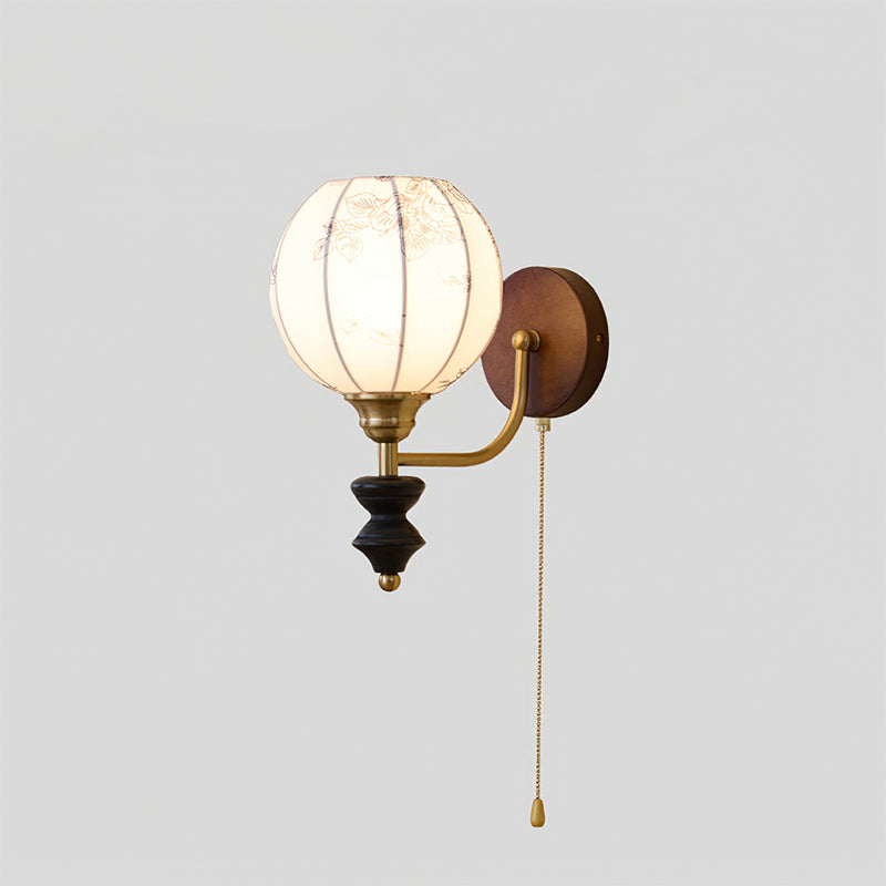 Clérachic wall light with a lit globe shade, a brass arm, a black wood pendant and a pull chain, on a plain grey backdrop.