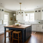 This Clérachic chandelier, in gold, hangs in a kitchen with white cabinets, an island with a wooden top and rattan stools.