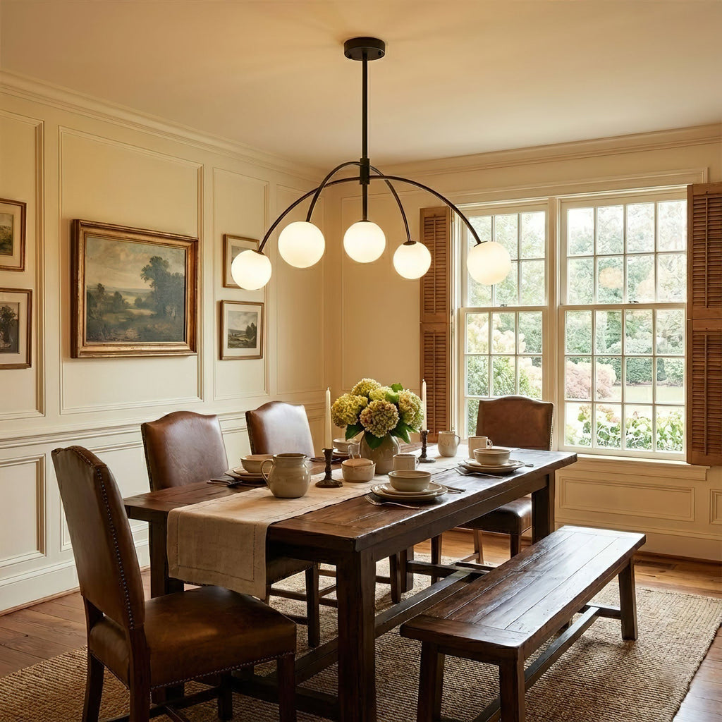 A black Clérachic chandelier hangs over a dark wood dining table with leather chairs and a bench, in a cream panelled room.
