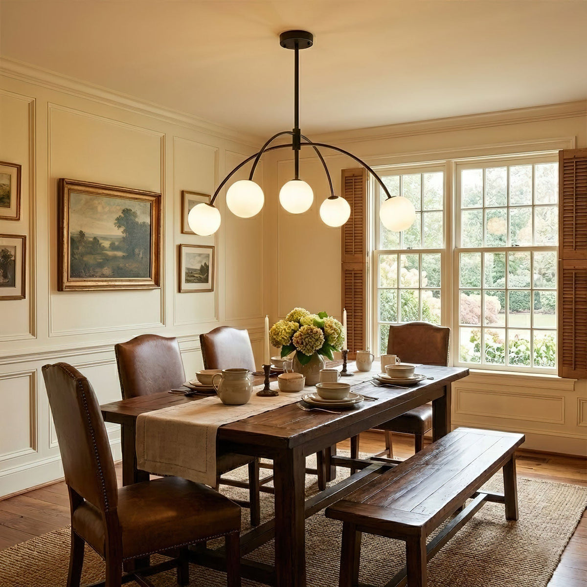 A black Clérachic chandelier hangs over a dark wood dining table with leather chairs and a bench, in a cream panelled room.