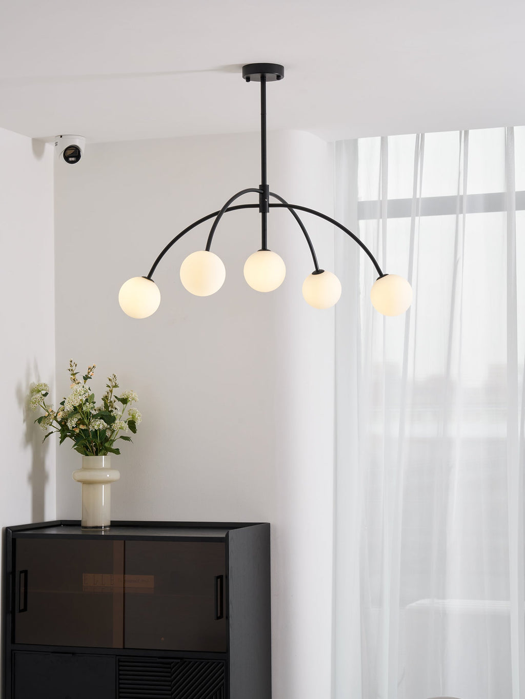 A black Clérachic chandelier with five lit globes hangs in a white room with sheer curtains, above a dark cabinet and a vase of flowers.