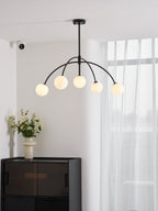 A black Clérachic chandelier with five lit globes hangs in a white room with sheer curtains, above a dark cabinet and a vase of flowers.