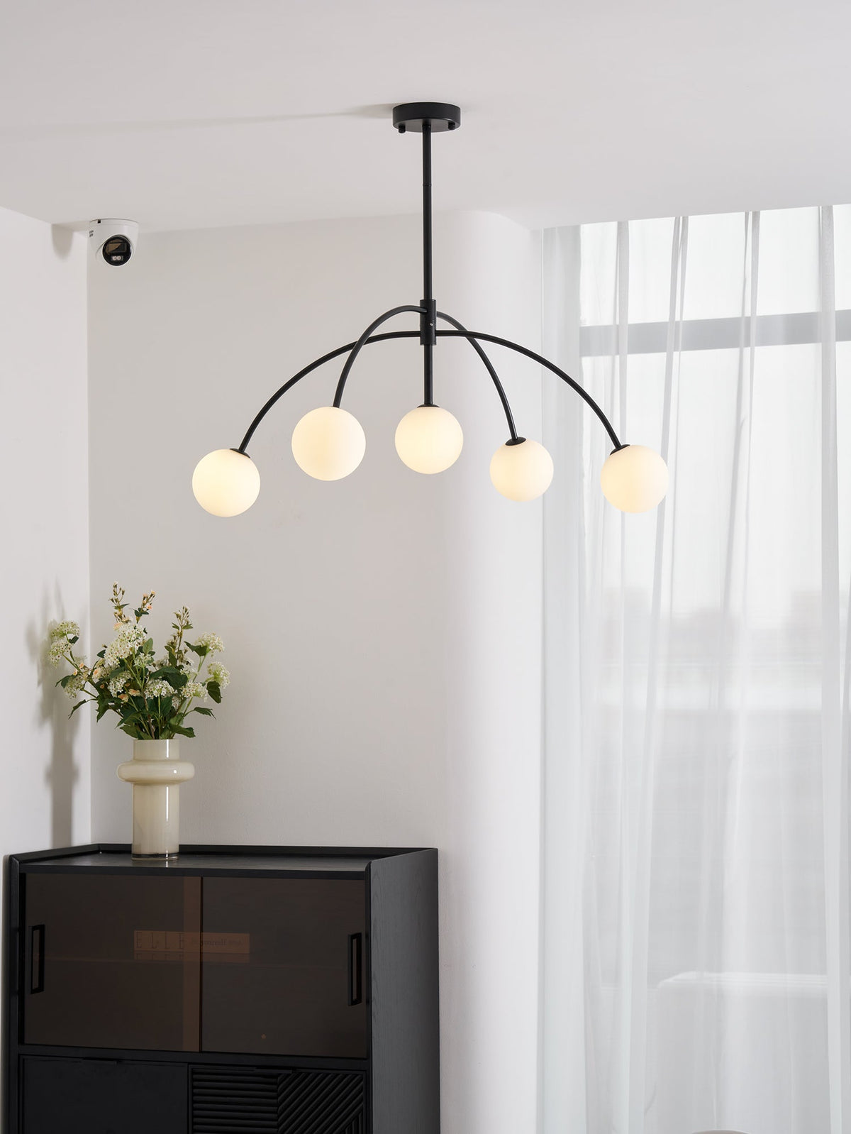 A black Clérachic chandelier with five lit globes hangs in a white room with sheer curtains, above a dark cabinet and a vase of flowers.