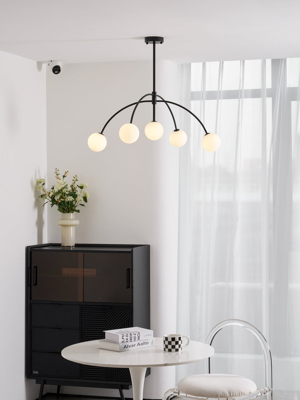 Five globes glow on this black Clérachic chandelier, which hangs above a round white table, a dark cabinet and a clear chair.