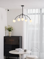 Five globes glow on this black Clérachic chandelier, which hangs above a round white table, a dark cabinet and a clear chair.