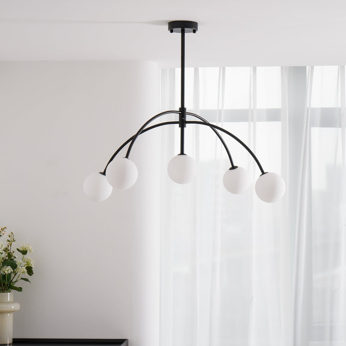 Slightly from below, a black Clérachic chandelier with five milky globes hangs against sheer white curtains, switched off.