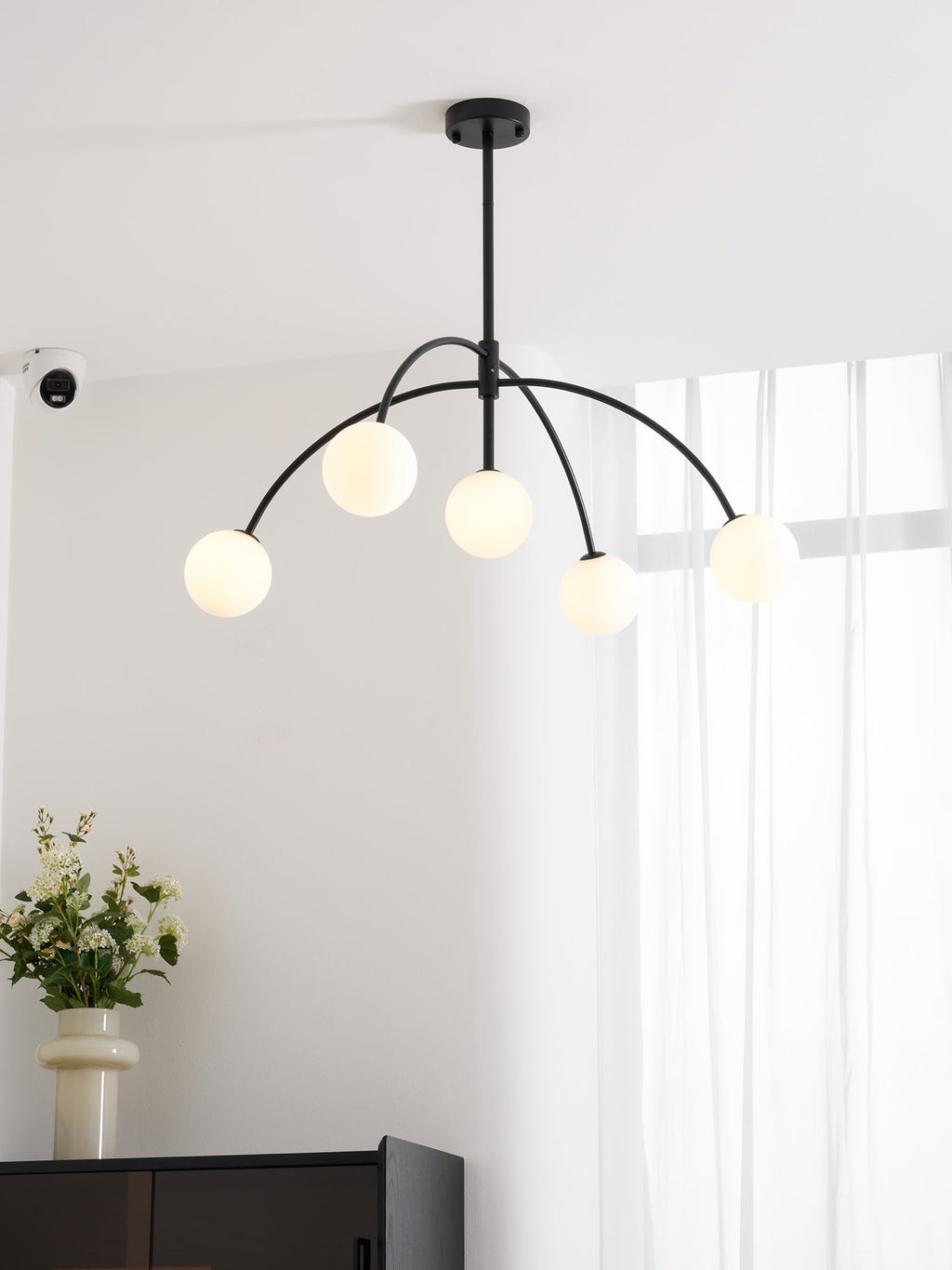 A black Clérachic chandelier with five lit globes hangs near a window, with a vase of white flowers and a dark cabinet below.