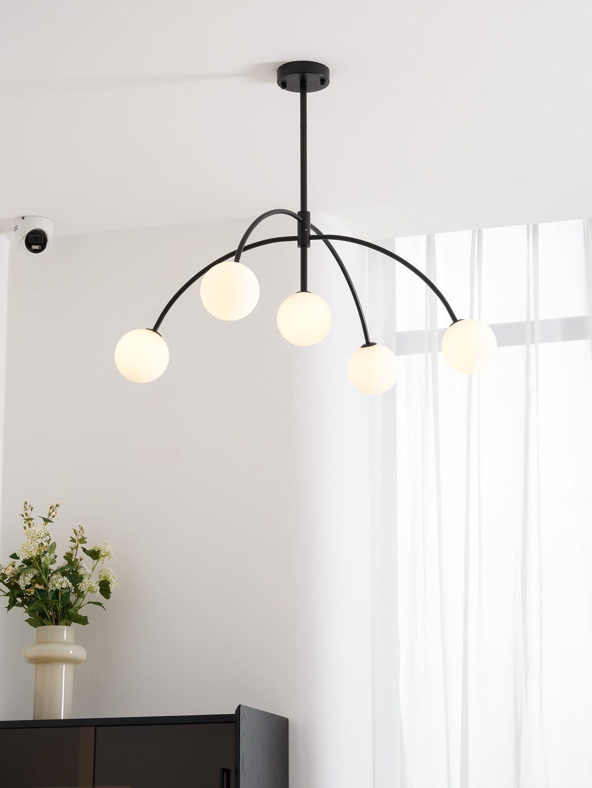 A black Clérachic chandelier with five lit globes hangs near a window, with a vase of white flowers and a dark cabinet below.