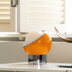 An amber-yellow version of this Clérachic table lamp sits on a white shelf by a window, its chrome rim tilted around a white dome.