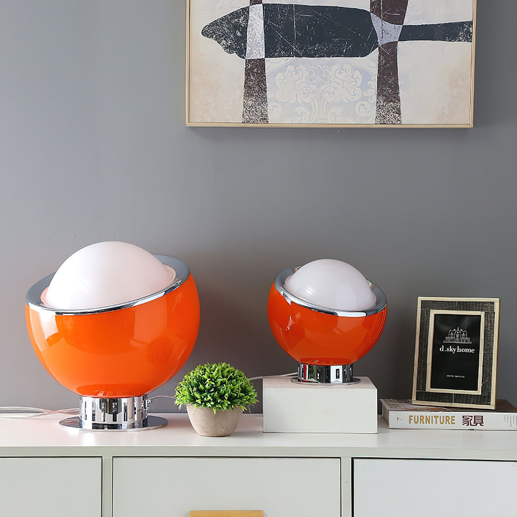 Two orange Clérachic table lamps, one large and one small, stand on a white sideboard beside a small plant, a framed print and a stack of books.