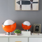 Two orange Clérachic table lamps, one large and one small, stand on a white sideboard beside a small plant, a framed print and a stack of books.