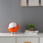 A single orange Clérachic table lamp sits on a white sideboard with a small potted plant, in front of a grey wall and a framed print.