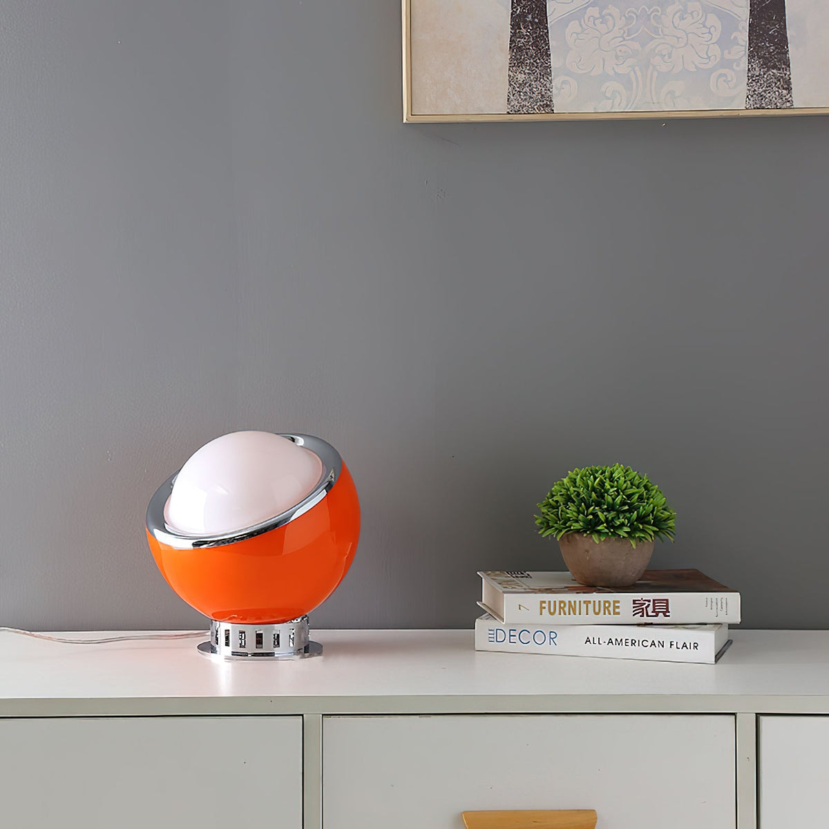 A single orange Clérachic table lamp sits on a white sideboard with a small potted plant, in front of a grey wall and a framed print.