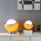 Two amber-yellow table lamps, one large and one small, glow on a white sideboard beside a plant and a framed picture, from Clérachic.