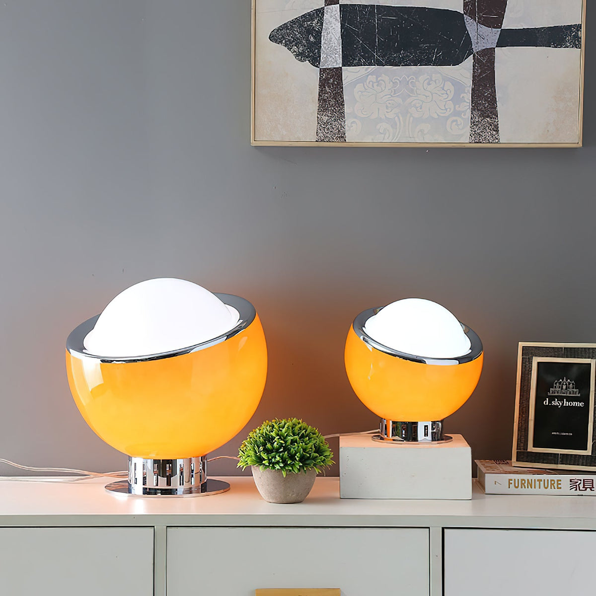 Two amber-yellow table lamps, one large and one small, glow on a white sideboard beside a plant and a framed picture, from Clérachic.
