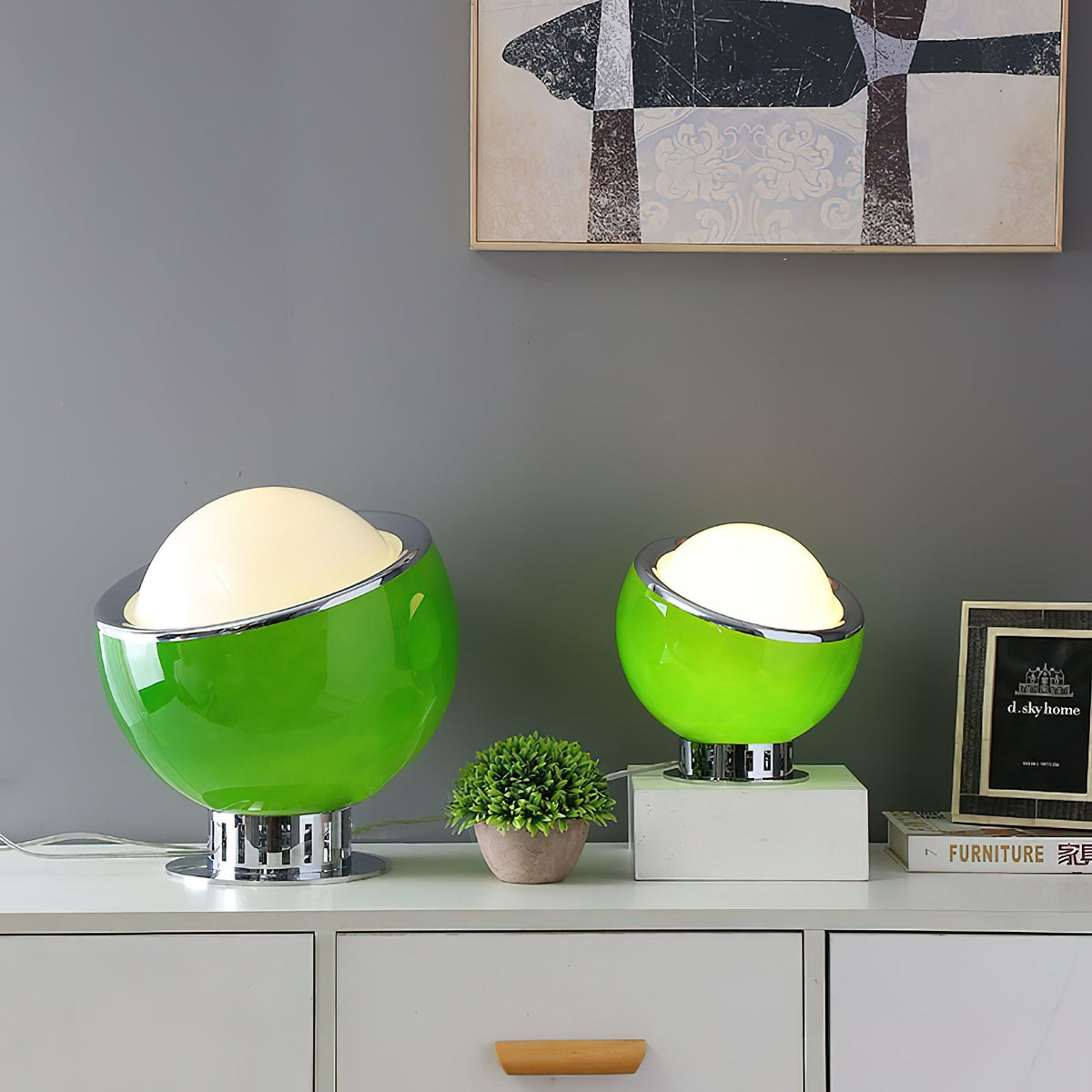 Green and lit, this Clérachic table lamp comes in two sizes on a white sideboard, beside a small plant and a framed picture.