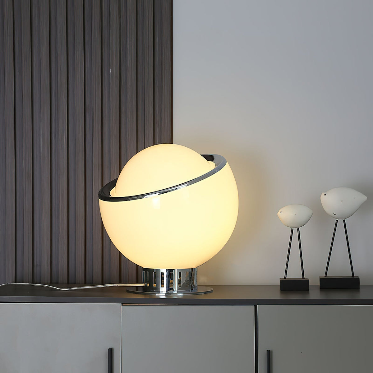 A lit white Clérachic table lamp glows on a dark sideboard in front of a slatted wall panel, next to two small bird figures on thin legs.