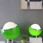 Two green Clérachic table lamps, large and small, sit on a white sideboard with a plant, the white domes lit against a grey wall.