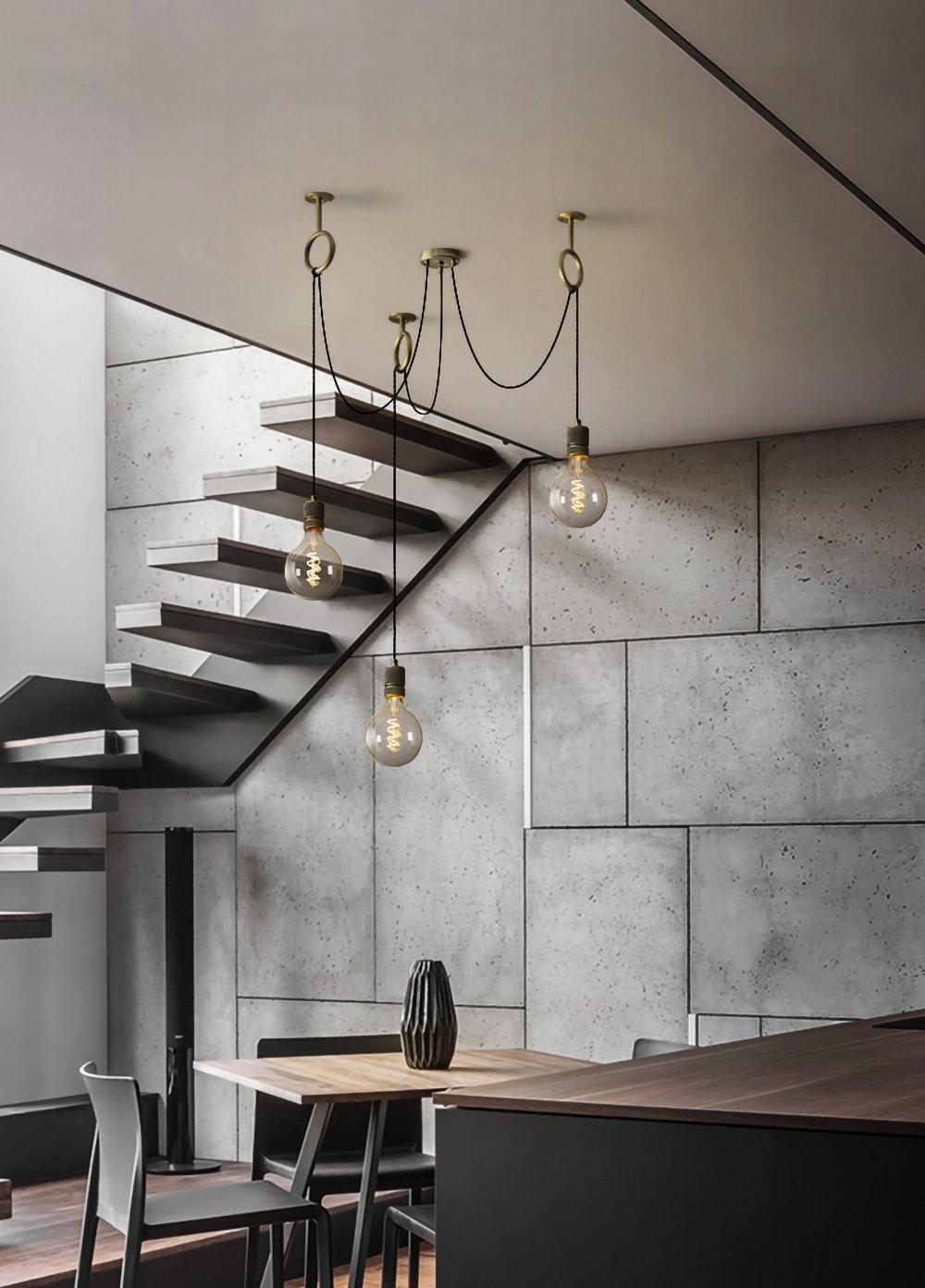 A small group of bulbs on this Clérachic pendant hangs beside a floating dark staircase and a grey concrete-tile wall.