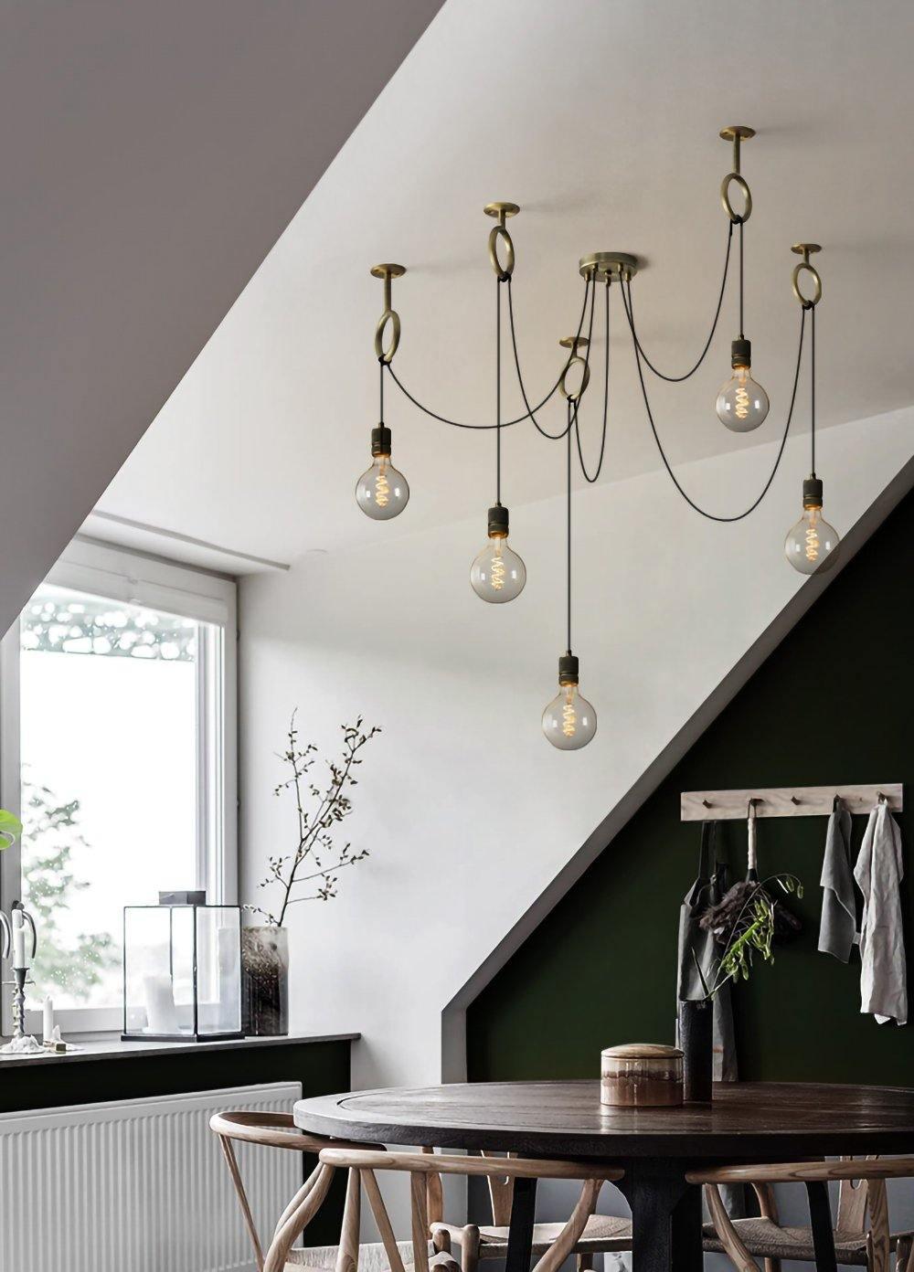 Globe bulbs of this Clérachic pendant hang beneath a sloped white ceiling near a dark green wall and a round dark dining table.