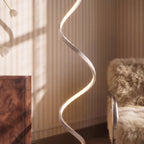 Clérachic spiral floor lamp close up, a pale satin ribbon with warm LED light along its edge, by a fur chair and a wooden cabinet.