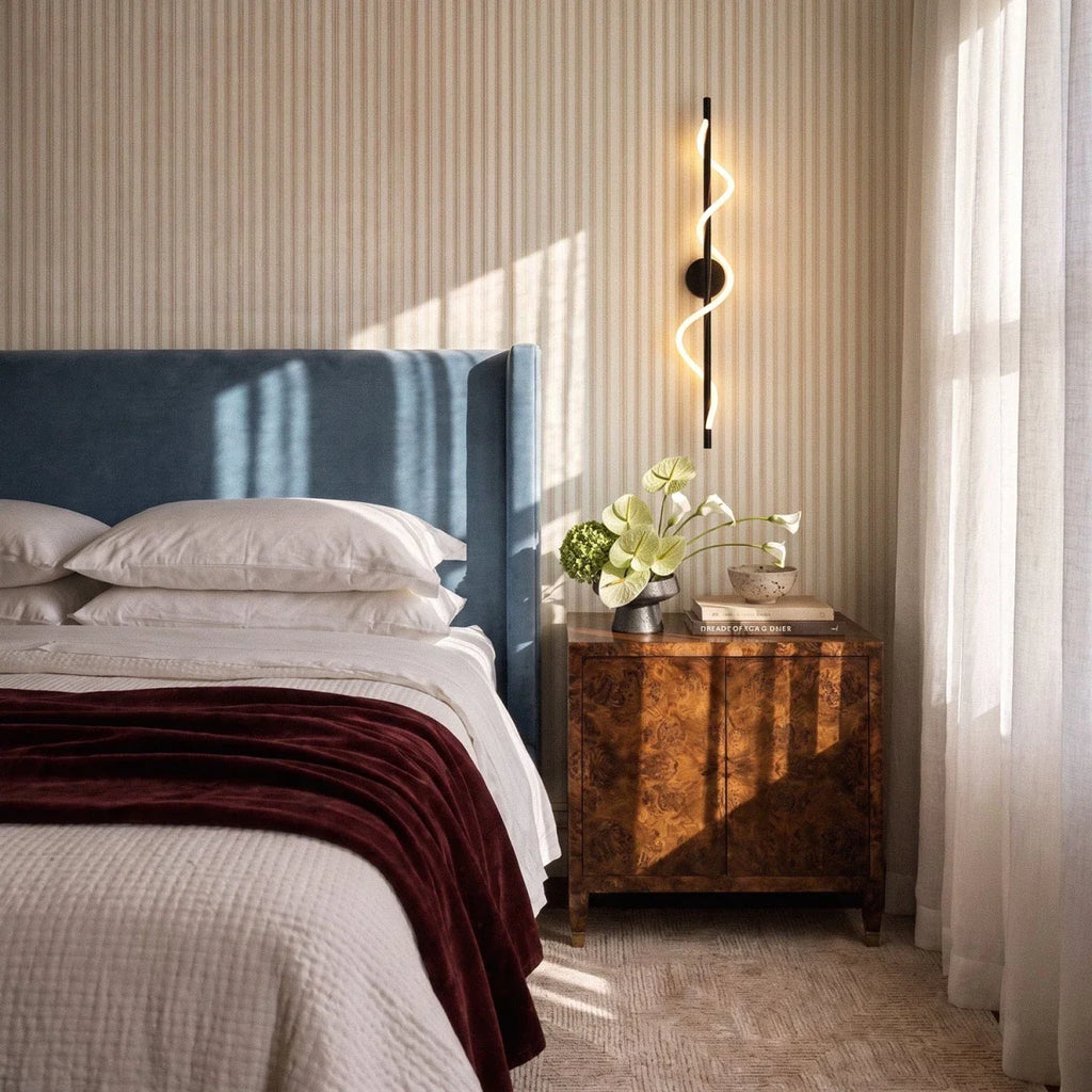Clérachic black spiral LED wall light glowing above a burr-wood bedside cabinet, beside a blue headboard and striped wall.