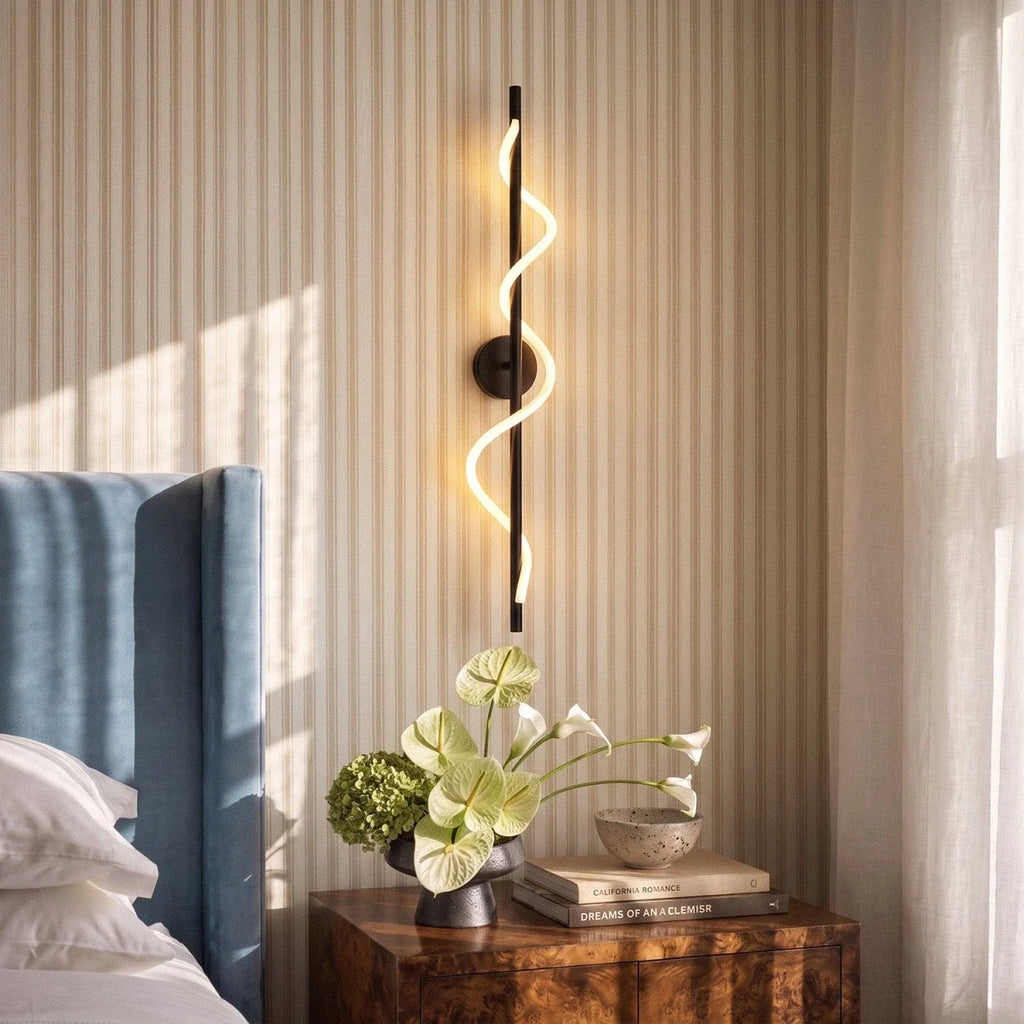 Clérachic black wall light with a glowing spiral LED strip over a bedside table with books, a bowl and green flowers.
