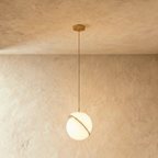 Clérachic larger opal white sphere pendant with a diagonal brass-finish ring, glowing from a brass rod below a plaster ceiling.