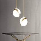 Two Clérachic opal white sphere pendants with brass-finish rings hung at different heights above a round marble table.