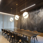 Three Clérachic opal white sphere pendants with brass-finish rings in graduated sizes above a long dining table and stone wall.