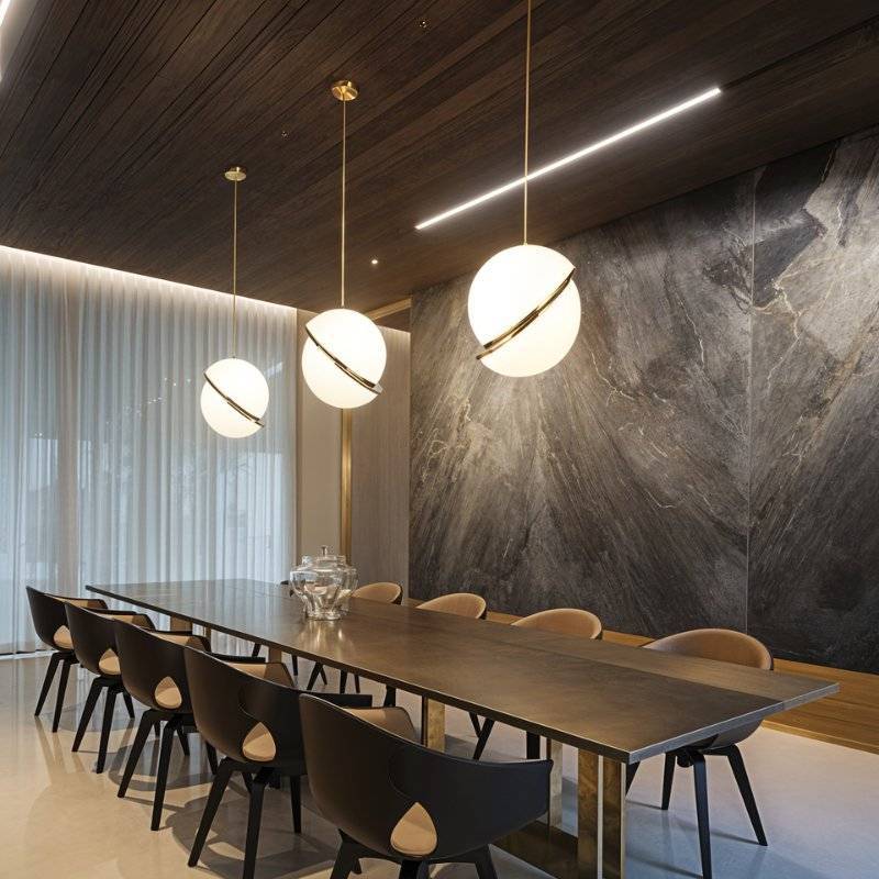 Three Clérachic opal white sphere pendants with brass-finish rings in graduated sizes above a long dining table and stone wall.