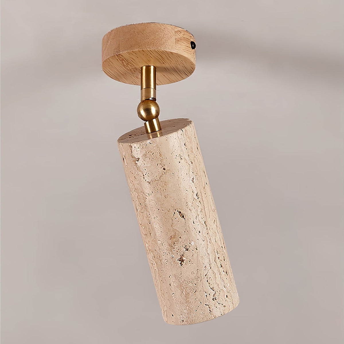 Close view of this Clérachic spotlight on a grey ceiling, with a pale wood canopy, a brass ball joint and a pitted travertine cylinder.