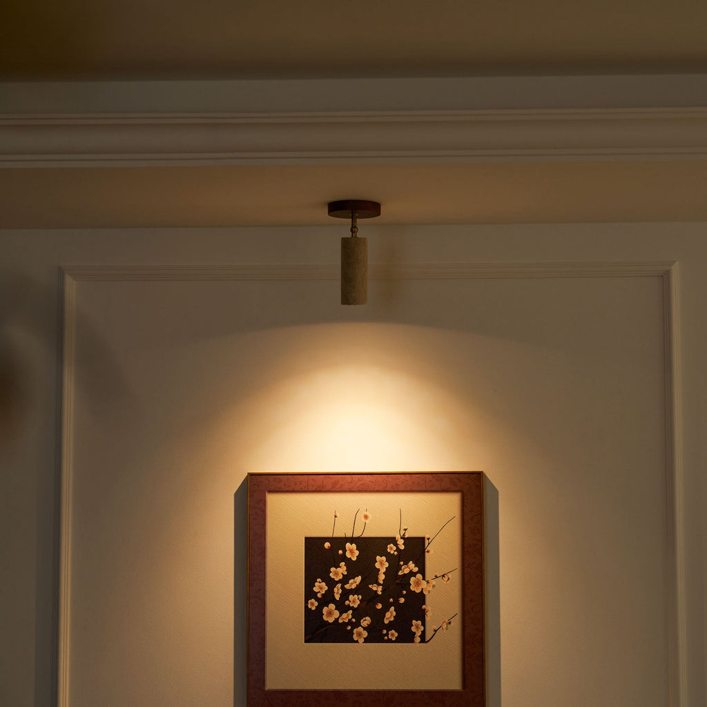 Lit Clérachic travertine spotlight in a dim room, casting a warm pool of light over a framed blossom print on a panelled wall.